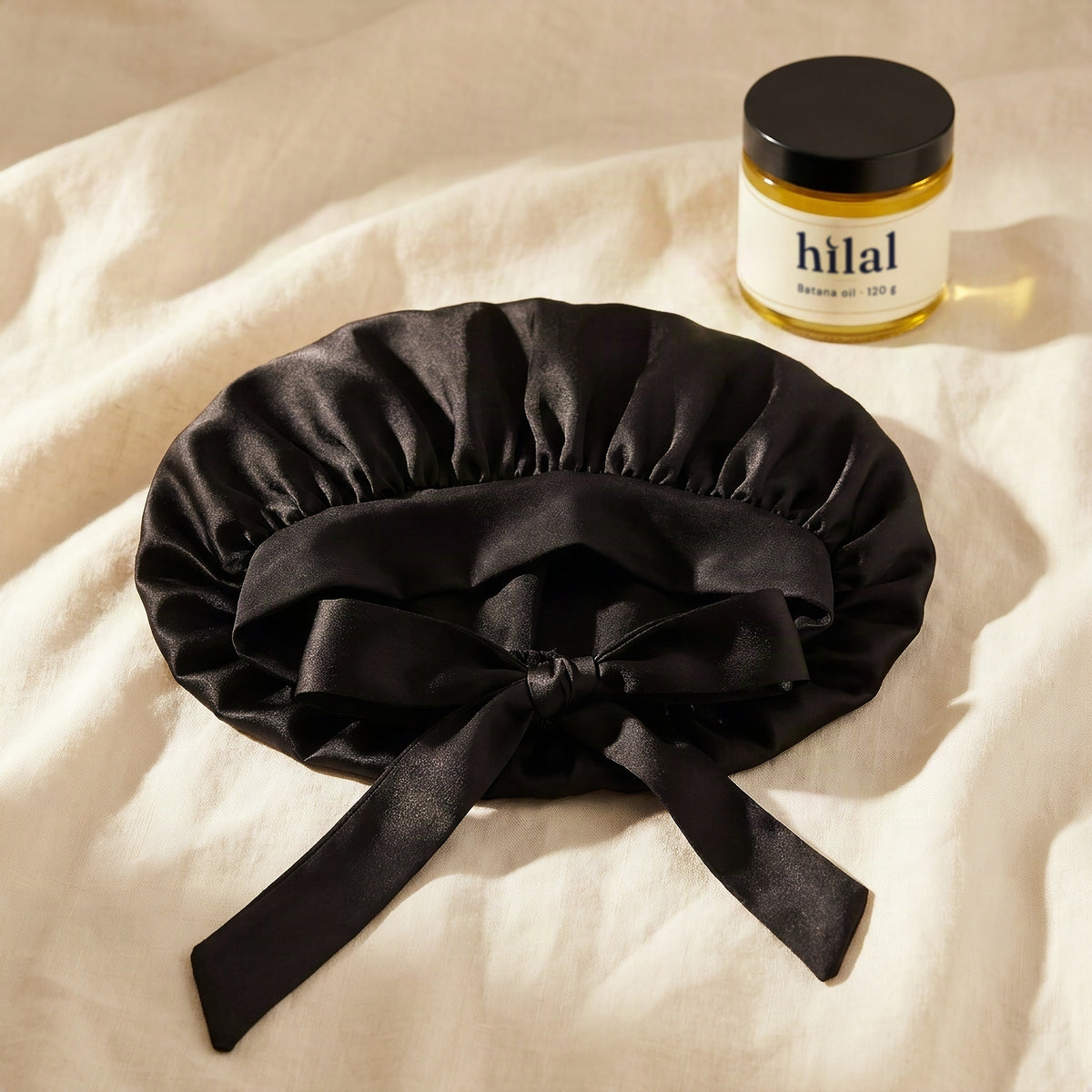 Satin bonnet on: the black satin bow bonnet on ivory linen with a jar of Hilal batana oil