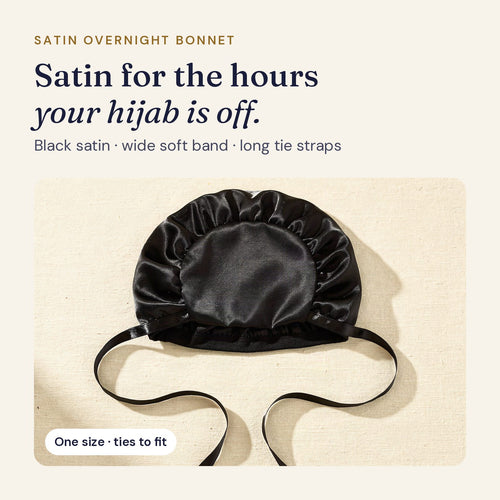 Black satin overnight bonnet with a wide soft band and long tie straps, laid flat on ivory linen
